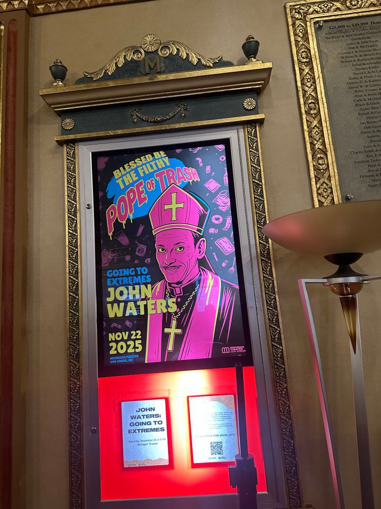 A photo of a poster on the wall. The poster—John Waters dressed like a pope in hot pink.