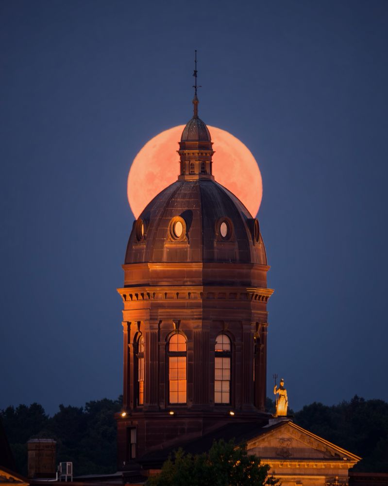 A large, orange full moon perfectly aligned with the rotunda atop the New Brunswick Legislative Assembly Building. The sky is a dull blue and the building is illuminated with warm lighting. 