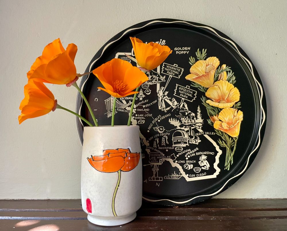 California poppies in a white ceramic vase with orange California poppies on it, in front of a black plate with a map of California and pictures of California poppies. 
