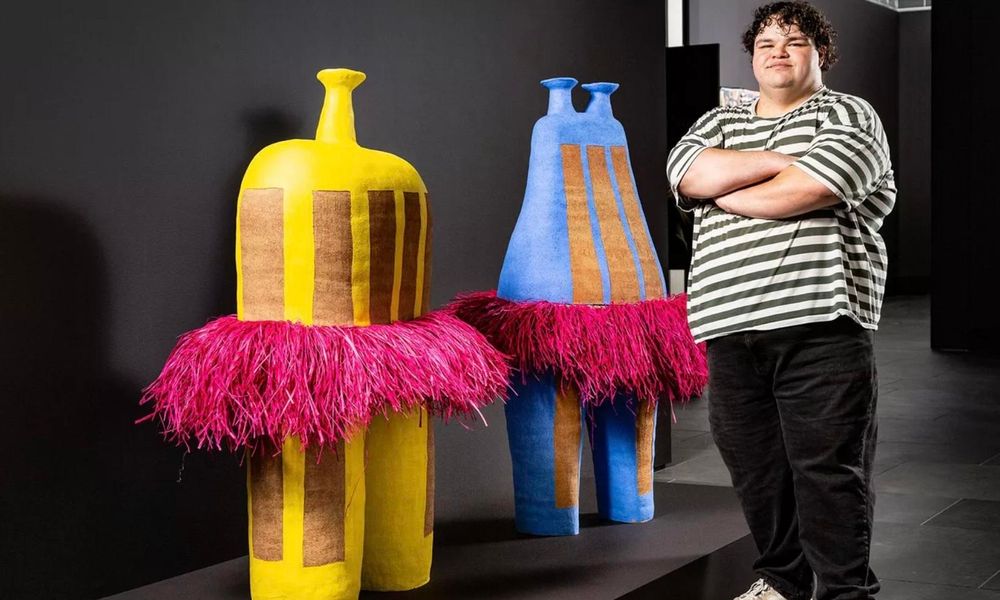 Alfred Lowe standing beside his bright yellow and blue overglazed brown ceramic works which are wearing pink raffia tutus.