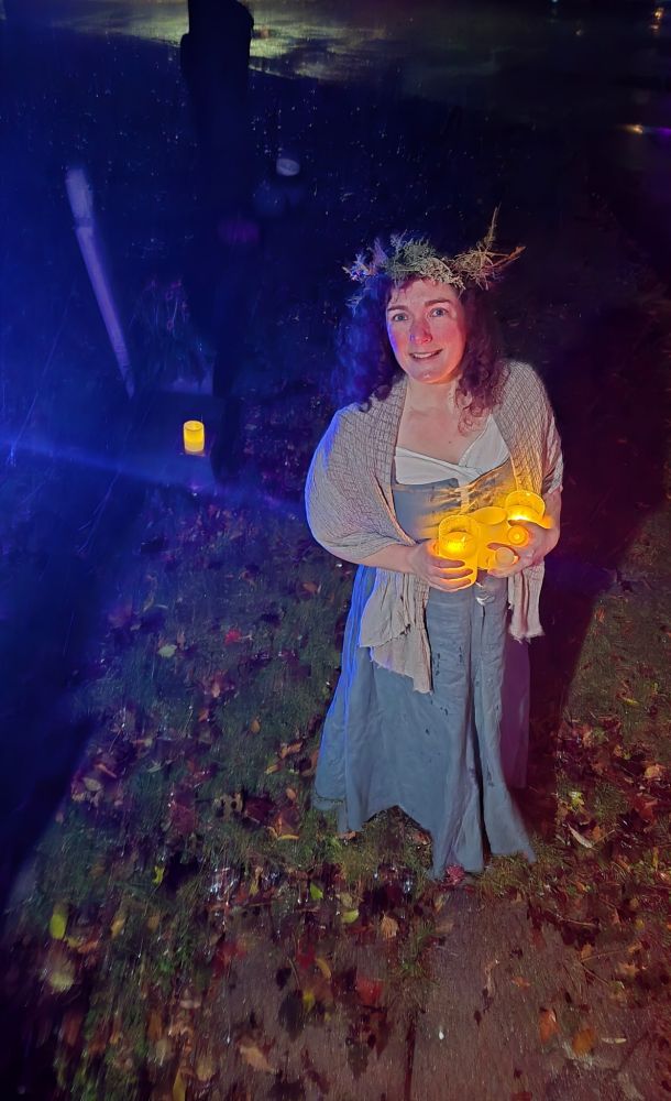 Acred faced lady in her green medieval dress and dried- leaf crown. She is bringing the fakey candles inside. Cold wet street and some Styrofoam tombstones behind her
