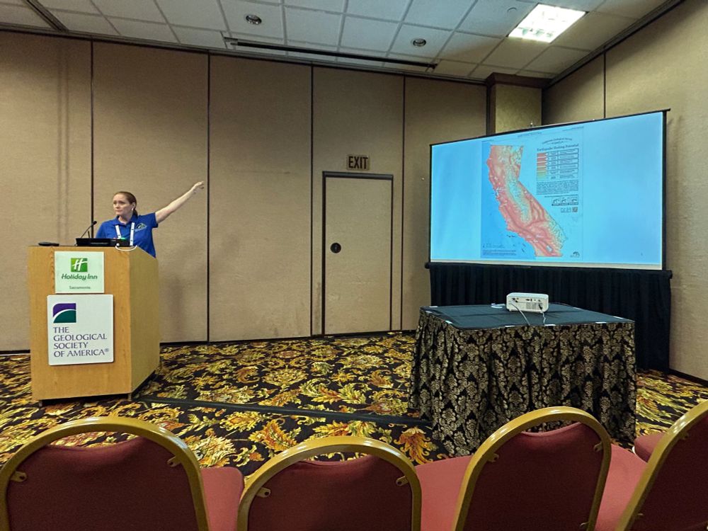 Photo of Wendy Bohon giving a presentation at the Geological Society of America, Cordilleran Section meeting in Sacramento. She is in a hotel conference room standing behind a wooden lectern with her arm lifted and extended towards the projection screen. On the screen is a map of California showing the largest potential earthquake shaking intensities for each area of the state. 