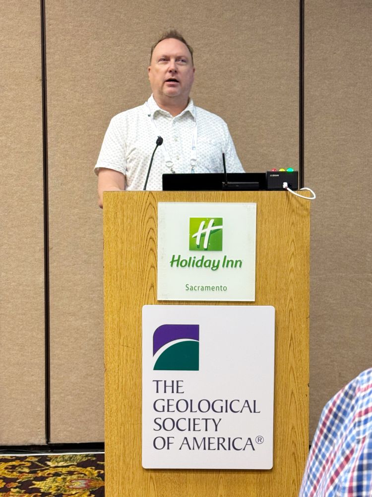 Photo of Brian Olson giving a presentation at the Geological Society of America, Cordilleran Section meeting in Sacramento. He is in a hotel conference room standing behind a wooden lectern addressing the audience. A sign at the top front of the lectern reads “Holiday Inn Sacramento” and another sign at the bottom reads “The Geological Society of America”.