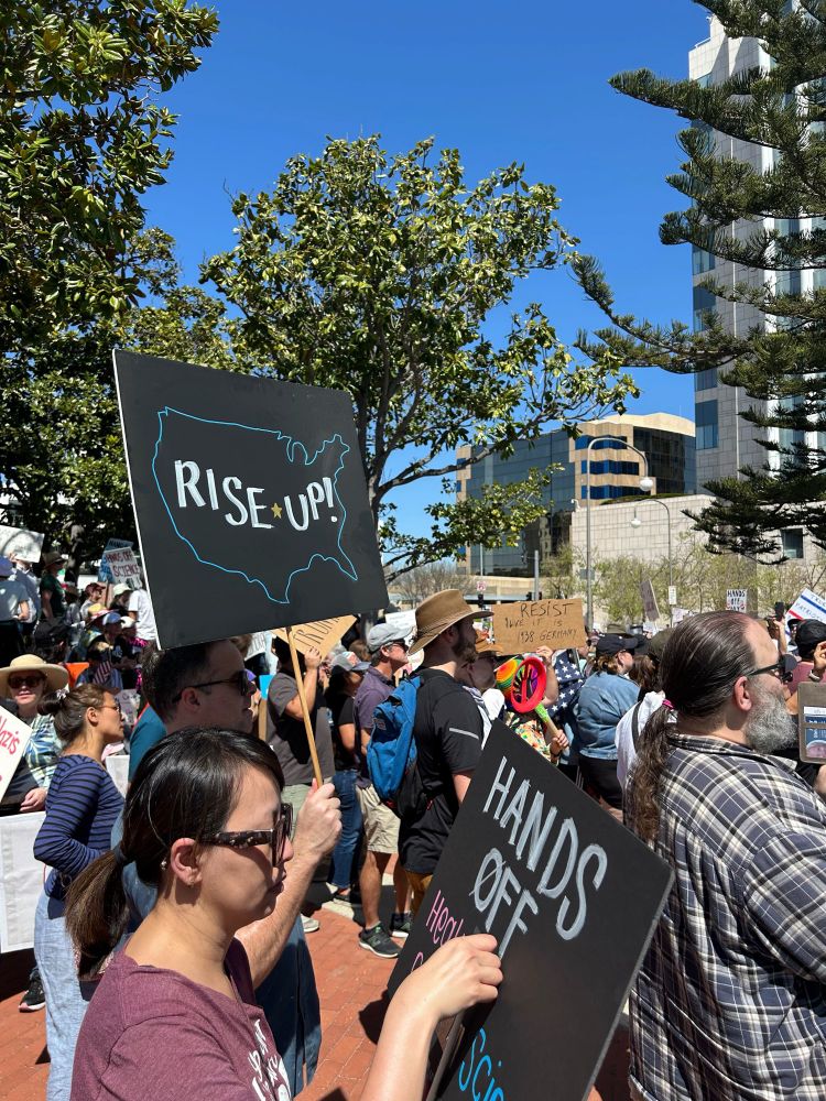 A crowd of protesters at a rally. One of them is holding up a black sign with an outline of the United States with the words “Rise Up!” written inside. 
