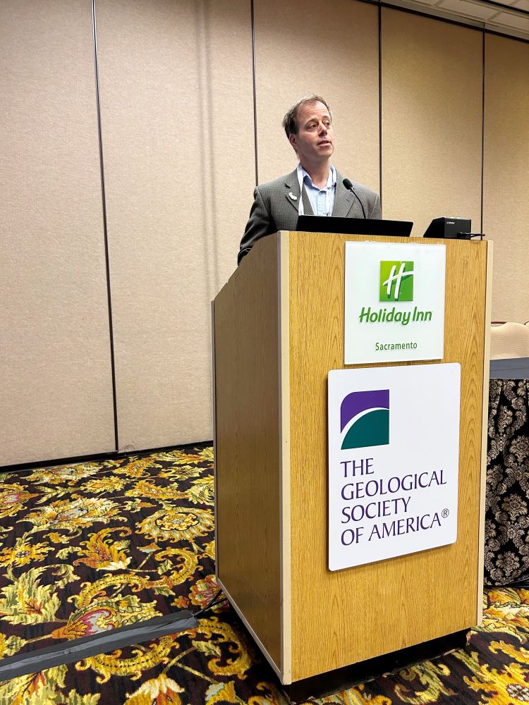California state geologist Jeremy Lancaster stands behind a podium speaking into the microphone giving a presentation. 