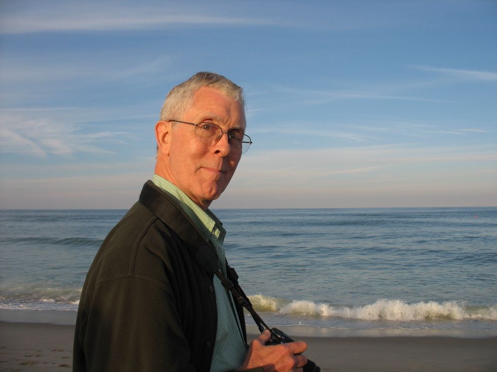 A man with grey hair and glasses, holding a camera around his neck and staring at the camera with a slight smile, while the ocean waves crash in the background 