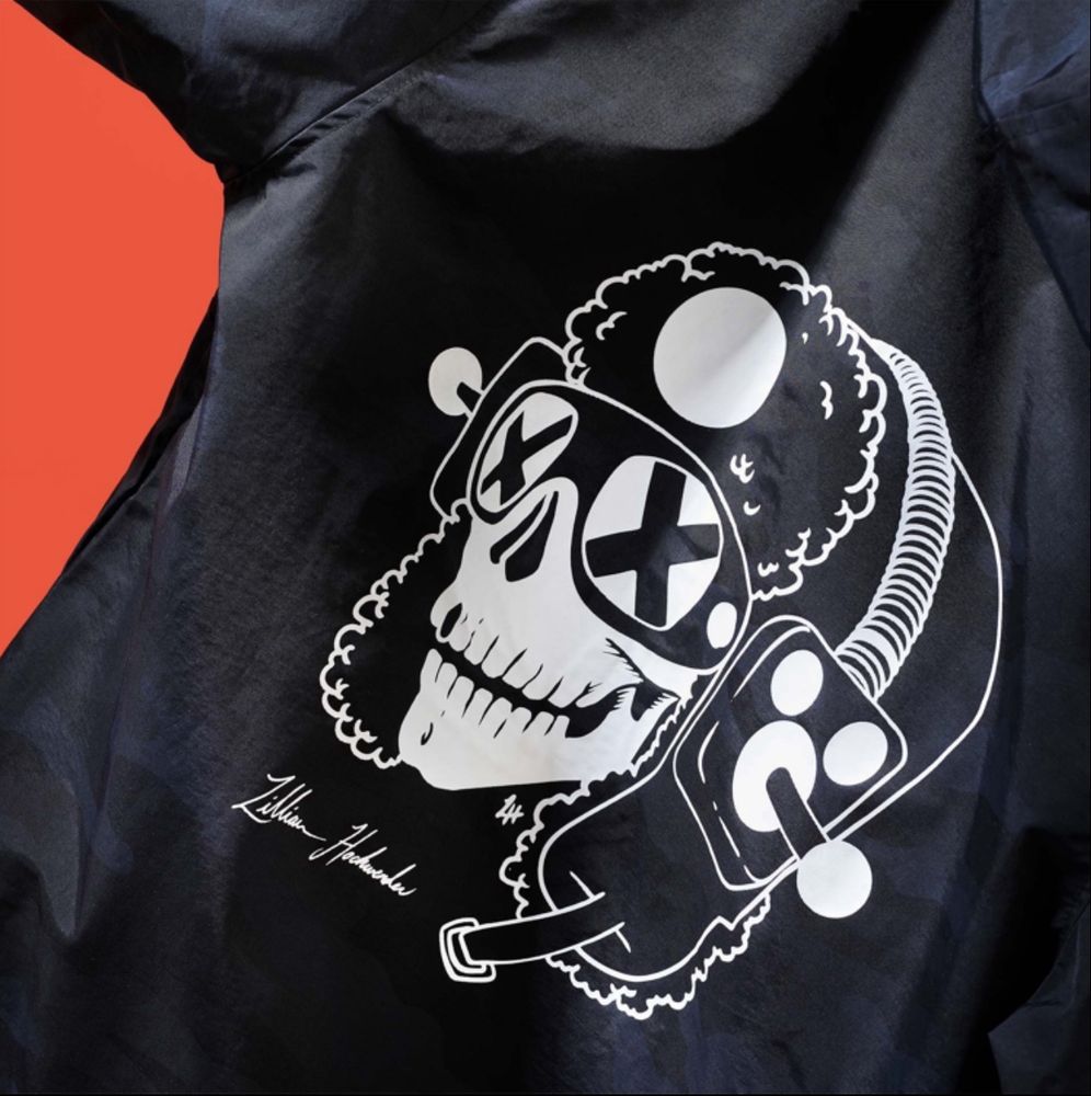 The back of a black jacket with a white line drawing of a skull in a furry aviator-style hat. The eyes of its ski goggles have crosses through them and it is wearing earmuffs over the hat which have joysticks where the puffs of the earmuffs would be. The illustrator's name (Lillian Hochwender) is signed underneath. The jacket is on a bright orange background.