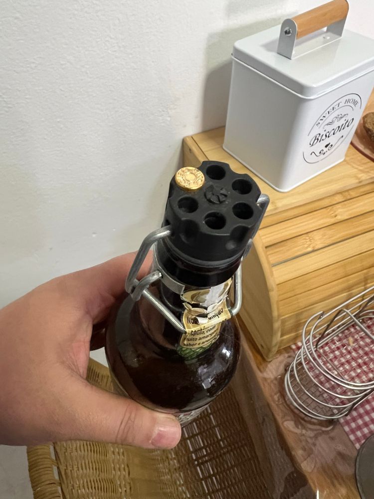 Um homem segurando uma garrafa de vidro no estilo belga. A tampa imita o barril de um revolver com apenas uma bala alocada no tambor, lembrando a famosa roleta russa.