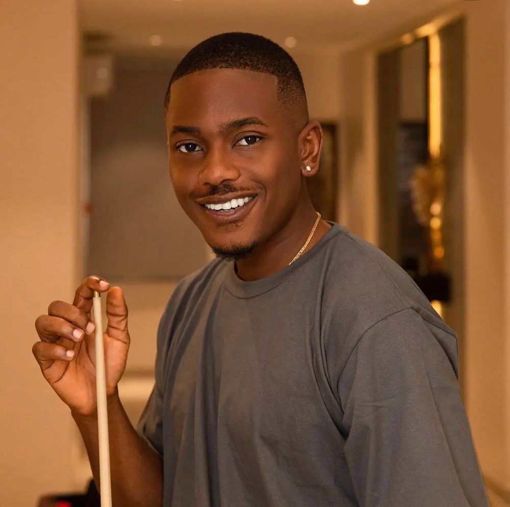 A young man with a charming smile standing inside a room, possibly a lounge. His hair has a clean fade leading to very short, almost buzzed hair. He wears a grey tee-shirt and thin gold necklace, tucked into the front of his shirt. He holds a pool stick, upright, as though it’s not his turn to shoot.