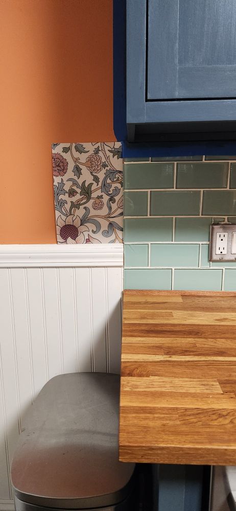 Close up of mountain blue cabinets, green glass tiles, butcher block counters, and a William Morris floral/paisley wallpaper with pink sunflowers, blue and green leaves, and yellow stems on an offwhite background.