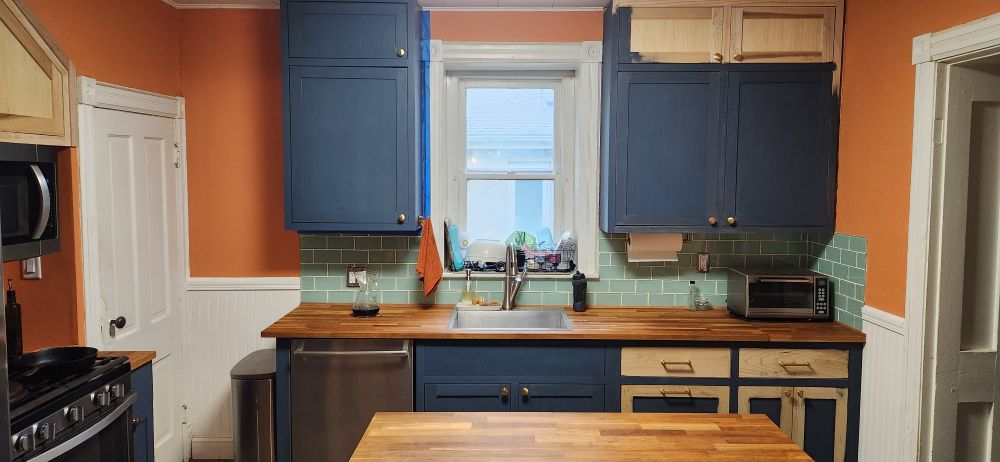 Wide angle view of partially painted shaker kitchen cabinets with brass pulls and knobs. Light green glass tile backsplash, and orange walls