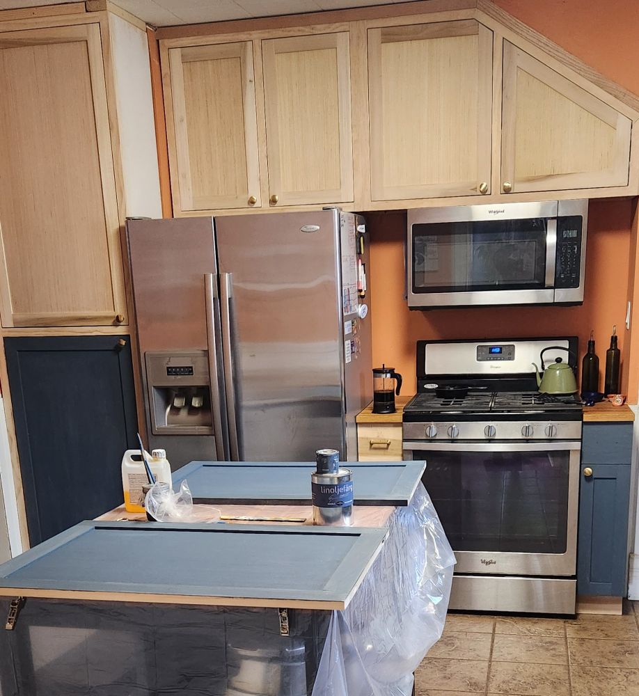 A kitchen with some cabinets painted blue and some raw wood, and the kitchen island is covered in drop cloth with two cabinet doors and linseed oil paint supplies on top.