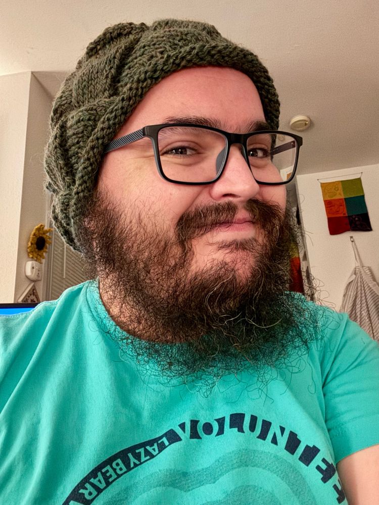 A smiling bearded person wearing glasses and a green knit cap looks into the camera