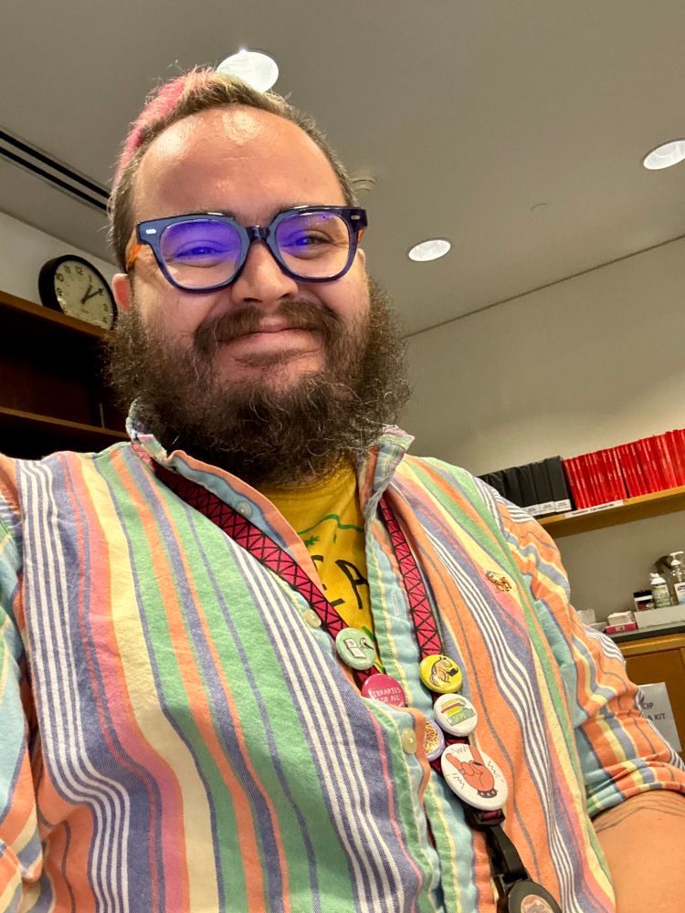 A bearded person with glasses smiles in tot he camera. They are wearing a colorfully striped shirt, and a lanyard with various buttons pinned on it. The background is generic office flavored