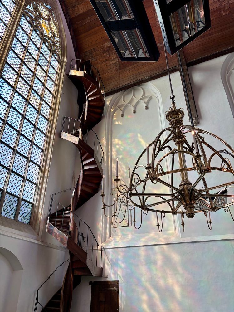 A tall siral staircase in the corner of a white room behind an ornate brass chandelier. A cathedral window on the left casts dappled and slightly chromatic light over the scene