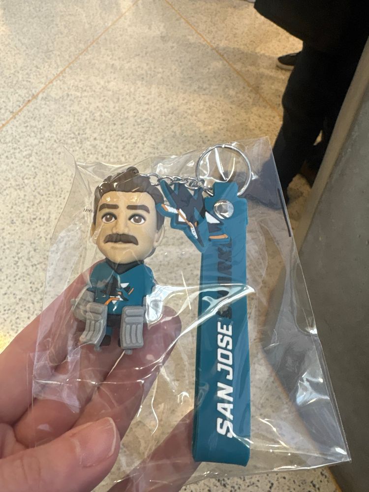 A keychain/bag tag with a teal strap that says San Jose Sharks, a small San Jose Sharks logo, and a chibi figurine of a hockey goalie in a San Jose Sharks jersey and gray pads, with dark hair and a mustache