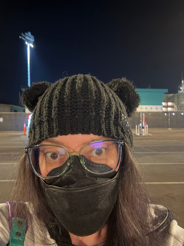 A brunette in a black beanie with double pompoms set to look like Mickey Mouse ears

She is also wearing glasses and a black mask