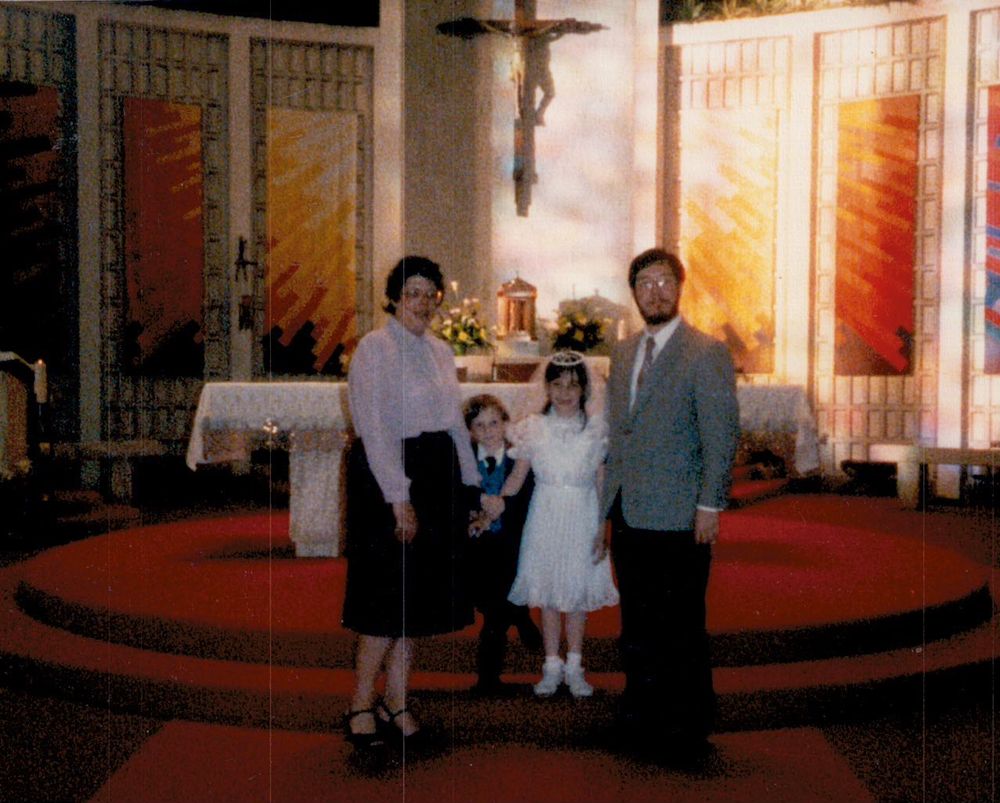 An 80s family standing in front of the altar in a Catholic Church. From the left, Mom, wearing a below-knee burgundy skirt, black Mary Jane heels, and a pale blouse; next is brother, wearing a dark suit with tie and blue sweater vest, clinging to the arms of Mom and sister respectively. Sister is wearing a ruffled white First Communion dress, small headdress with veil, and white Mary Janes with ankle socks, while Dad is wearing a grey sport coat with dark slacks. The family visually frames the golden tabernacle behind the altar with the crucifix above it, and sunburst banners hang on the walls to either side of the crucifix.