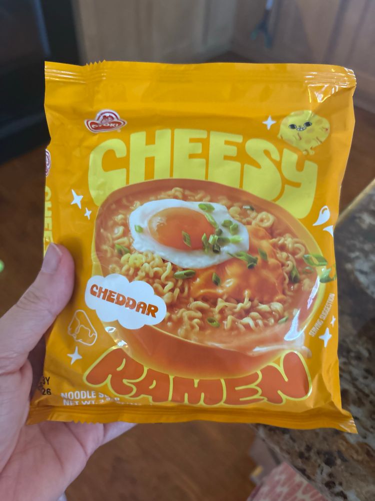 A packet of Otoki Cheesy Cheddar Ramen. The packet is bright orange-yellow with a picture of a bowl of ramen with a melted slice of cheese, a fried egg and sliced green onions on top, with a cartoon cheese character on the top right corner of the packet