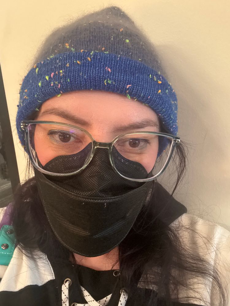 A dark haired woman wearing blue glasses, a blue and gray beanie, and a color blocked white and black hoodie with lacing at the neckline like a hockey sweater. She looks tired and is about to purchase a huckleberry cider, which will not help with the tiredness but will be delicious.