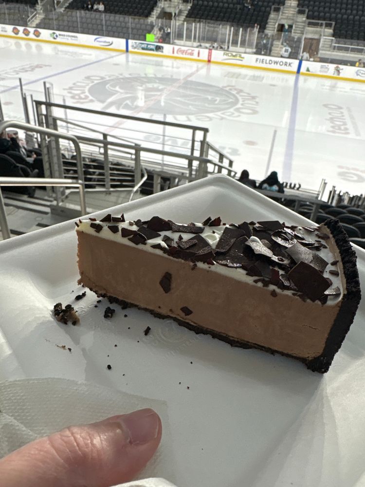 A slice of chocolate mousse cake on a square paper plate, with the Tech CU Arena ice sheet in the background