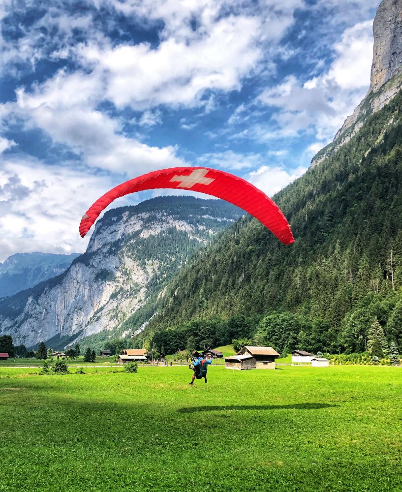 The photo depicts a paragliding scene in a mountainous valley. A person is captured mid-air, suspended beneath a large, red paraglider canopy featuring a white cross, the Swiss flag emblem. The paraglider is landing in a green meadow, with mountains in the background.