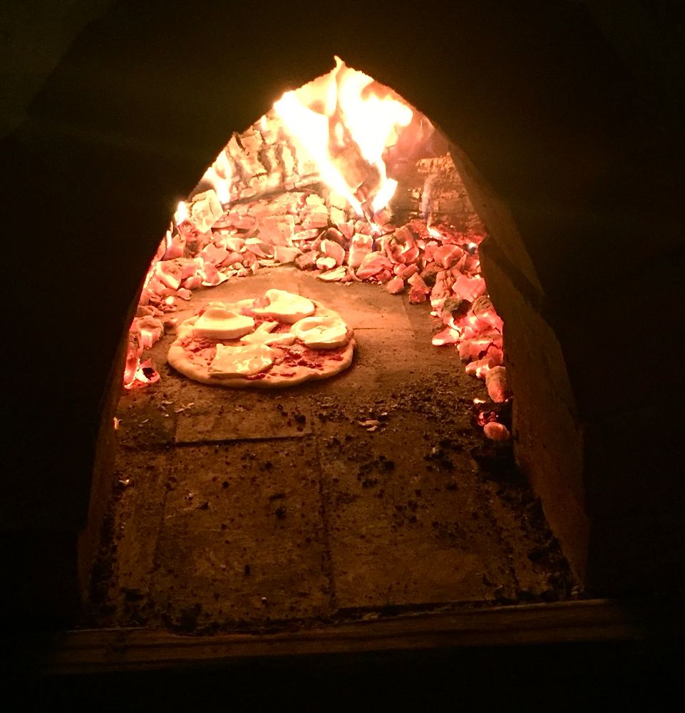 The photo depicts a pizza cooking inside a wood-fired oven. The oven's interior is illuminated by vibrant flames and glowing embers. The pizza, topped with cheese and other ingredients, rests on the oven floor.