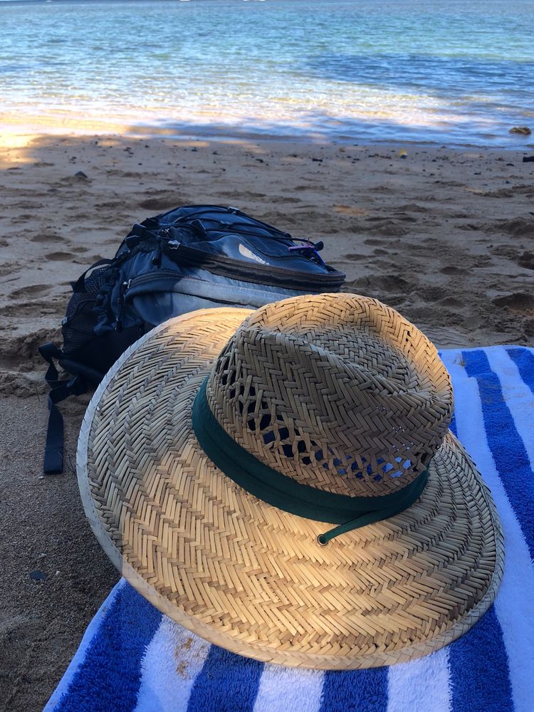 The image depicts a tranquil beach scene, focusing on a straw sun hat with a dark green band resting on a blue and white striped towel in the foreground. Behind the towel and hat, a dark-colored backpack is visible on the sandy beach. The calm ocean is in the background.