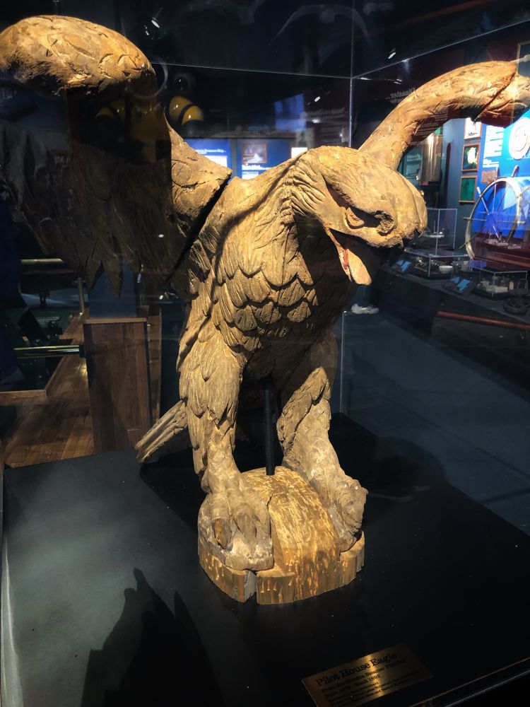 The photo captures a large, carved wooden eagle statue, displayed within a glass case in a museum. The eagle is posed with outstretched wings. The wood is weathered, and it appears that the beak is missing.