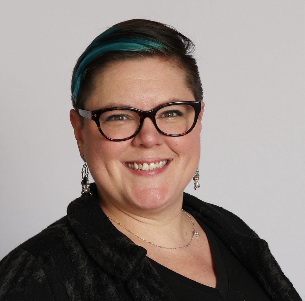 A professional headshot of me. I am wearing a black blazer, tortoise shell glasses, dangly black earrings and I have blue hair 