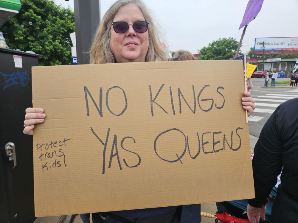 Woman in sunglasses (me!) with “No Kings Yas Queens” protest sign. It says “Protect trans kids” in small print. 
