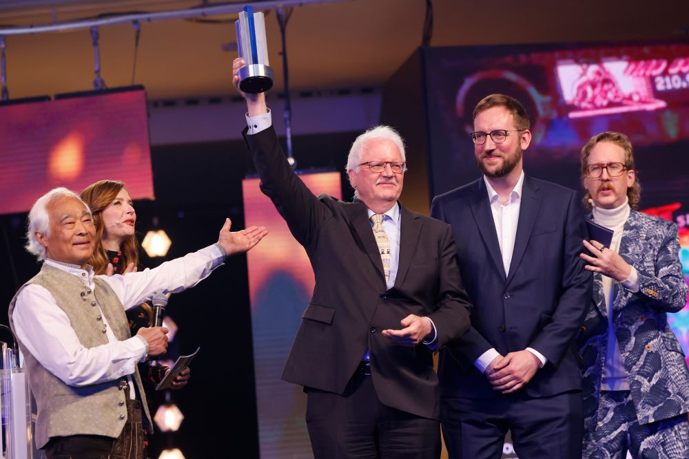 Rainer Krapohl vom FAMS reckt auf der Bühne des DCP 2025 die DCP-Trophäe empor. Neben ihm steht Steffen Bergmann ebenfalls vom FAMS. Zudem auf dem Bild sind die Moderator*innen Katrin Bauerfeind und Uke Bosse, sowie der Laudator Takeo Ischi.
