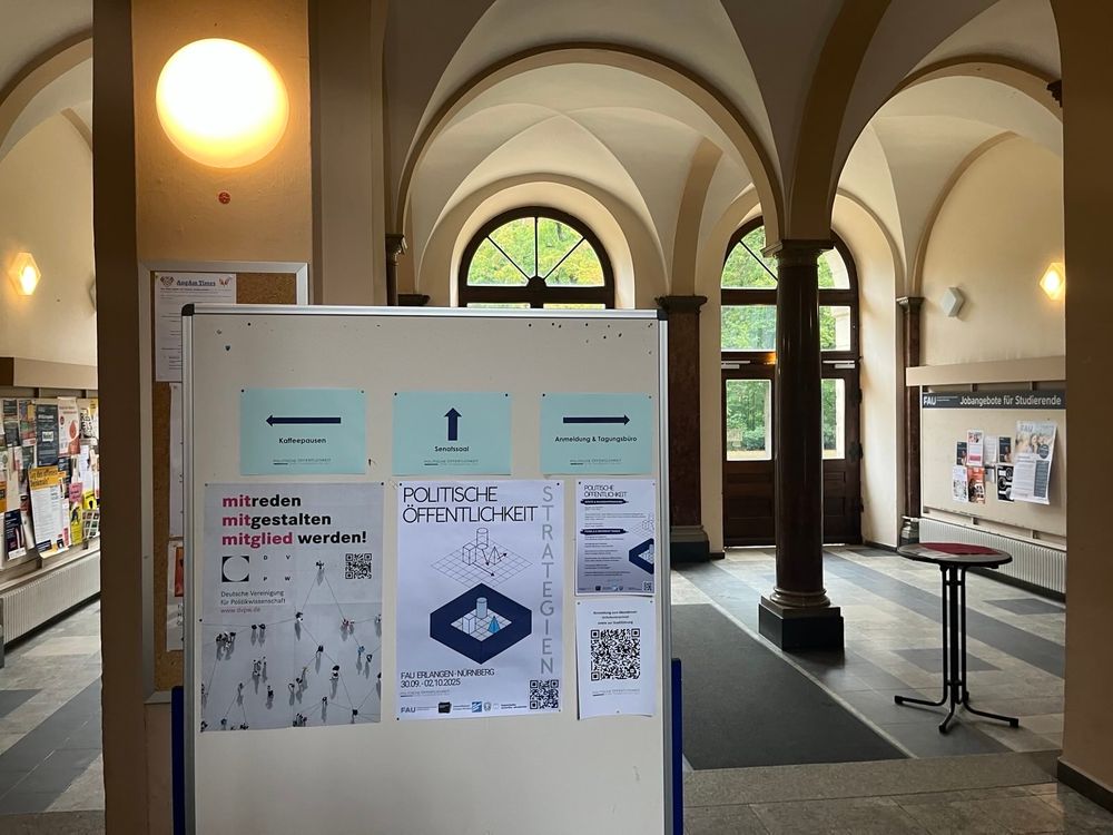 Man sieht die Eingangshalle des Kollegienhauses mit einer Infotafel unserer Tagung, auf der Wegweiser und Plakate hängen. 