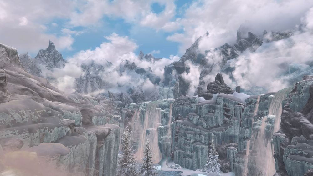 A screenshot of the Forgotten Vale from Skyrim during a bright day. Waterfalls tumble over glaciers into a half frozen lake as clouds wrap around the mountains in the background.
