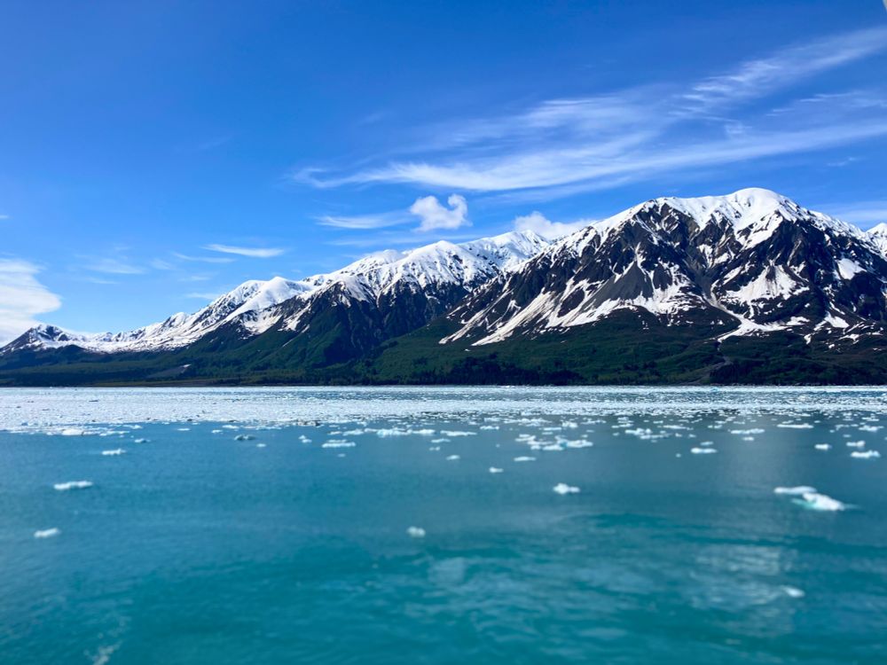 Snow-capped mountain range under a bright blue sky with wispy clouds and one is the shape of a small animal like a squirrel or chipmunk. Icy turquoise water and floating ice chunks in the foreground.