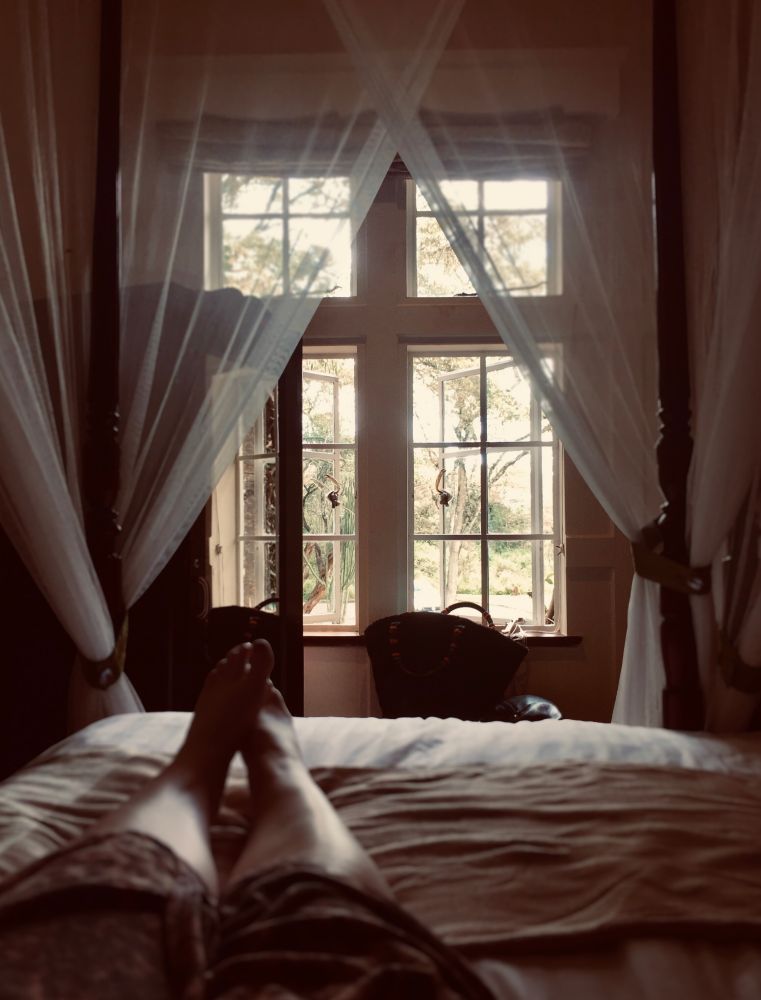 a woman on a bed with mosquito netting tied to the posts. The afternoon light in the room is fading and the view out the open window shows a gentle light at the end of the day through the trees.