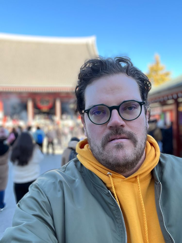 Me a man in a yellow hoodie and green jacket looking displeased at a temple. 