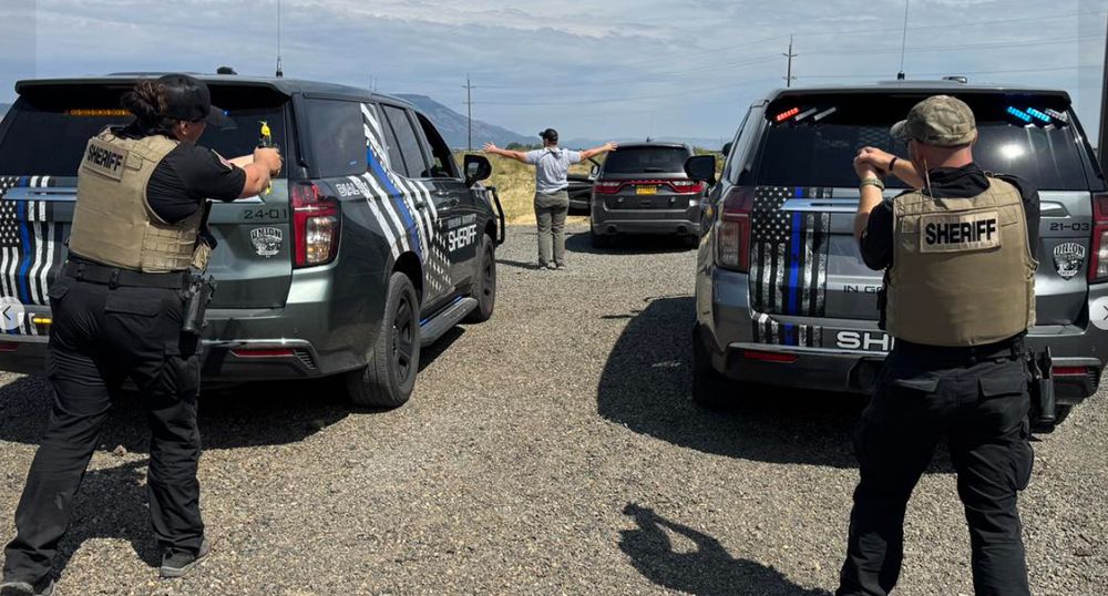Image of 2 sheriff deputies with guns drawn on a man. Two police SUVs draped in black and white US flags with a thin blue line strip and the words "In God We Trust." 