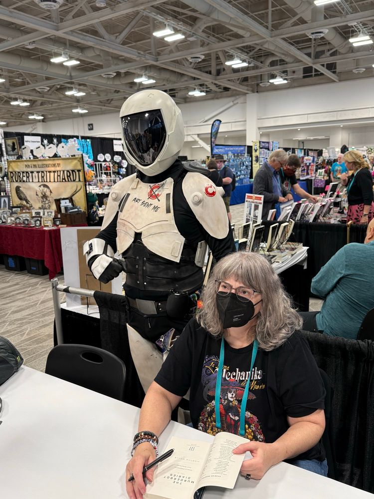 Murderbot stands behind Ms Wells as she autographs books at World Con. 