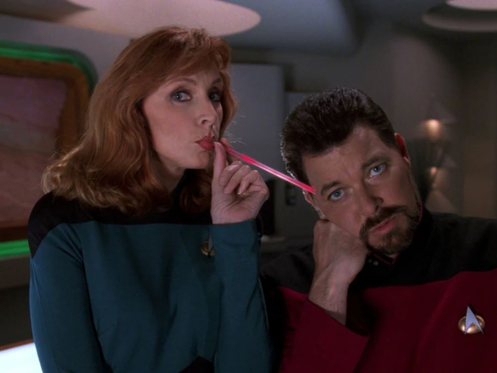 Dr. Crusher in Ten Forward sucking from a pink straw connected to Riker's forehead. They both look quizzically at Data, off-camera.