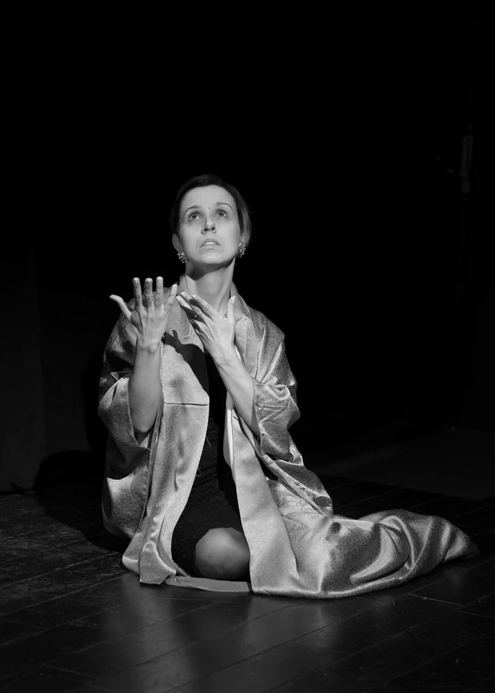Photo en noir et blanc prise par Soriya Thach.
Jarm0u en costume de Praskovia (robe noire de veuve, robe de chambre longue en satin) est agenouillée dans une pose de veuve en douleur, le regard vers le ciel.