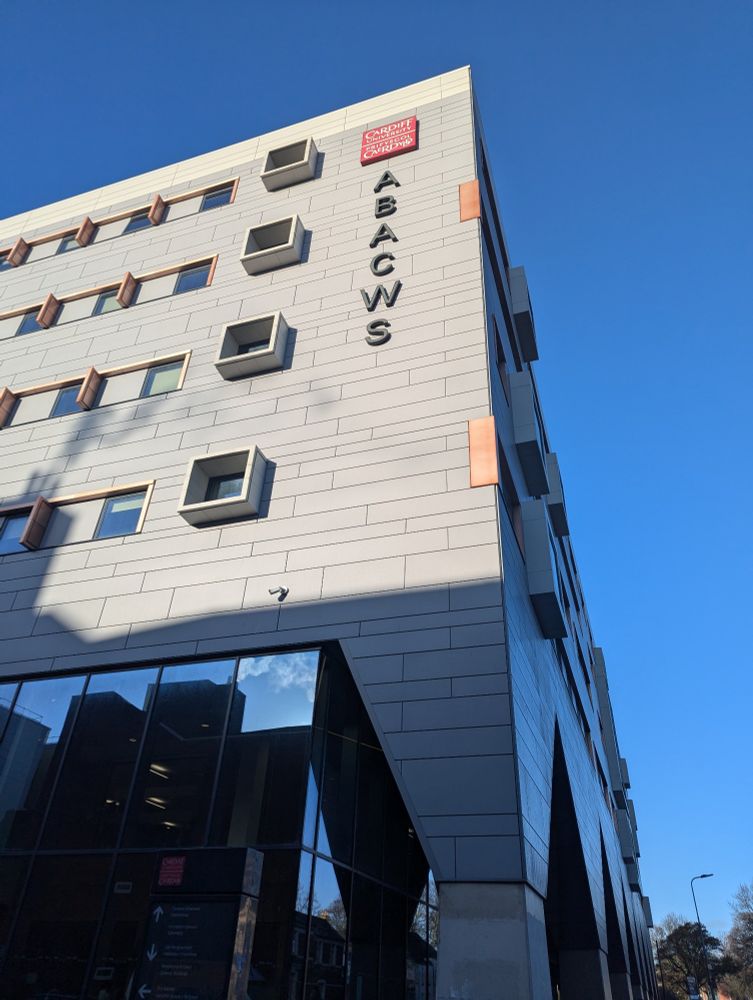 The maths department building at Cardiff. The Cardiff university logo - a red square with "Cardiff University, Prifysgol Caerdydd" at the top of the building with the word "ABACWS", the name of the building, going vertically down the side. The view is leaning up from the base of the building with a blue sky behind it. The building is modern with slate grey mismatched panelling, copper window frames, and the ground floor has tall glass walls.