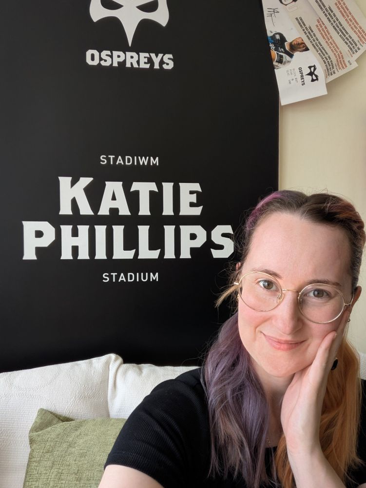 A selfie of Kat sat infront of a plaquard that has the ospreys logo and the text "Ospreys, Stadiwm Katie Phillips Stadium" written on it. Kat is smiling at the camera with her head resting slightly in her open palm. She has split orange and purple medium length hair which is pinned back, and is wearing a plain black tshirt and has round golden glasses. 