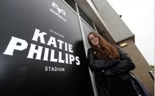 A young Kat stands next to a plaquard that has "Ospreys, Stadiwm Katie Phillips Stadium" on it. the plaquard is part of the branding attached to the wall of the Stadium. Kat has long brown hair and glasses, and is leaning on the wall with her arms crossed. She's wearing a leather jacket and smiling down at the camera. 