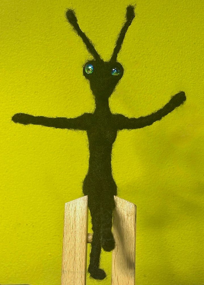 needle felted bug/figure sits on top of a mini easel awaiting her wings.  needle felted black yarn and loose roving fibers over wire with iridecent black glass bead eyes.