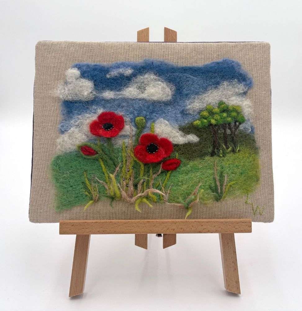 Foreground Poppies (2025) - needle and wet felt (background) wool painting by Lynn Wartski. Bright right red poppies in forground of image with rollong hills under a blue sky with fair weather clouds and small copse of trees to the side. Seed bead embellishment in flower centers and among blades of grass.