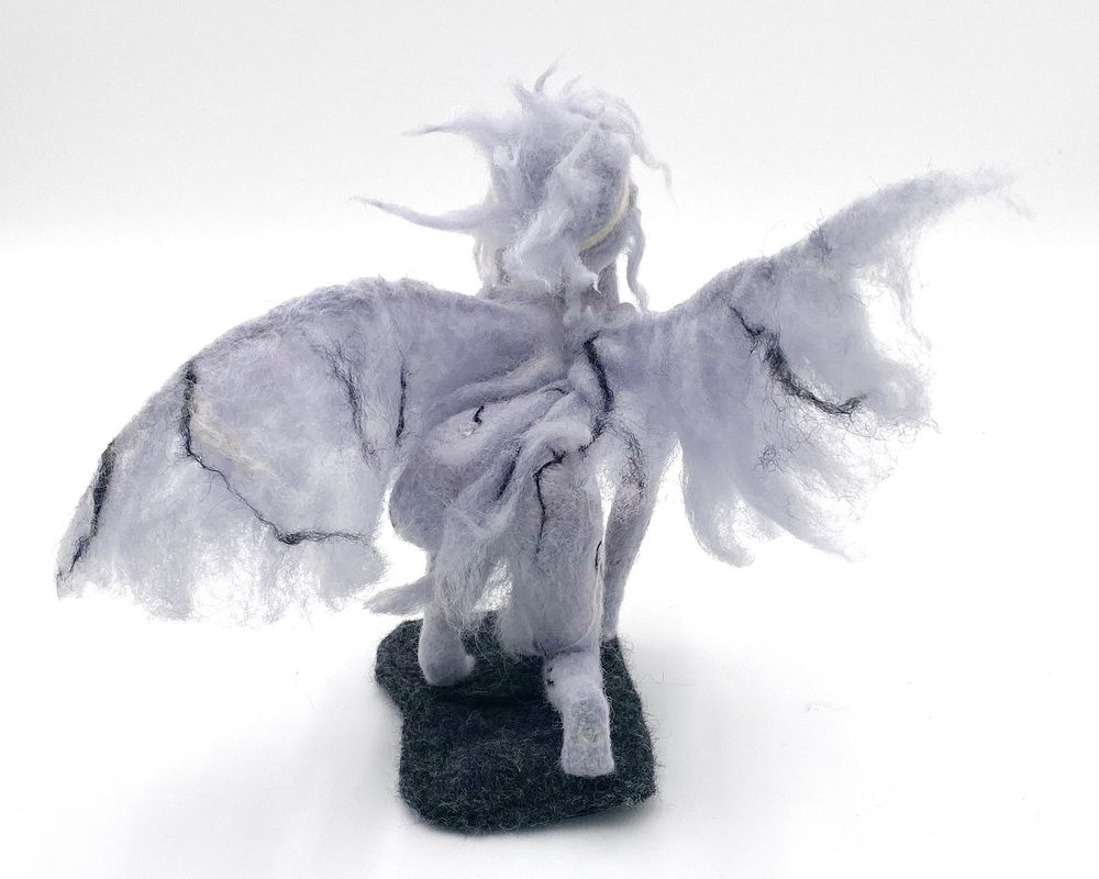 Back view of "Rise" needle felted figure sculpture of a rising young winged liberty by Lynn Wartski.  Grey veined wool mimics marble surface of a statue, iridecent galss bead eyes, a "bloodied" bandage wraps forehead, and her diadem crown is grasped firmly in her left hand. Figure's hair is loose, and her clothes and wings are tattered