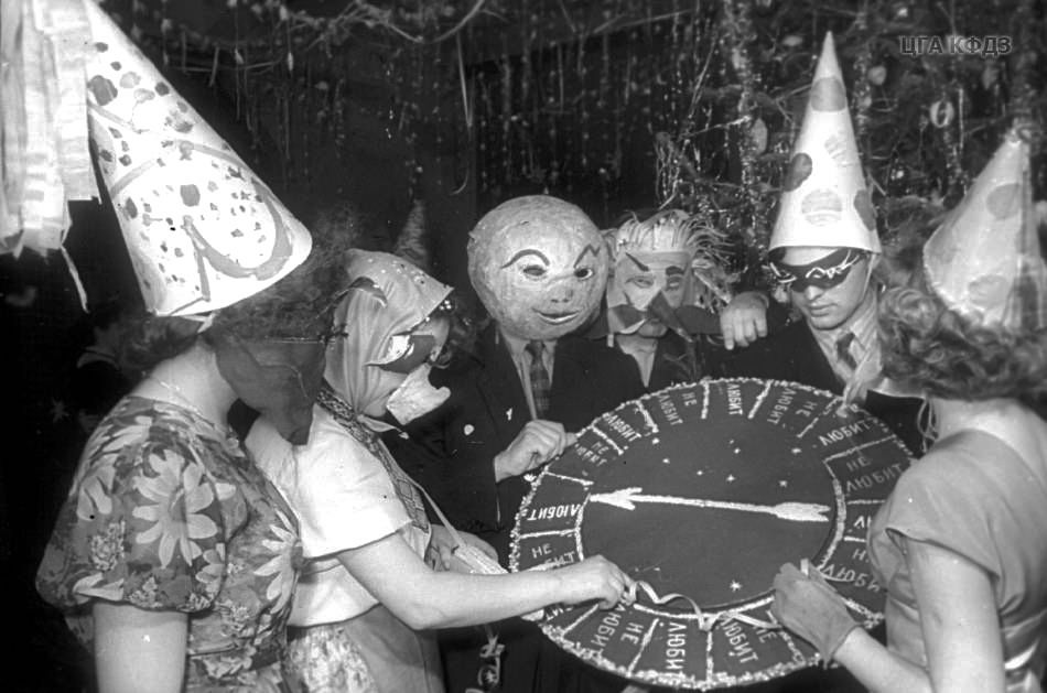 A vintage photo of a group of people in fancy dress and quirky masks spinning a fortune telling wheel.