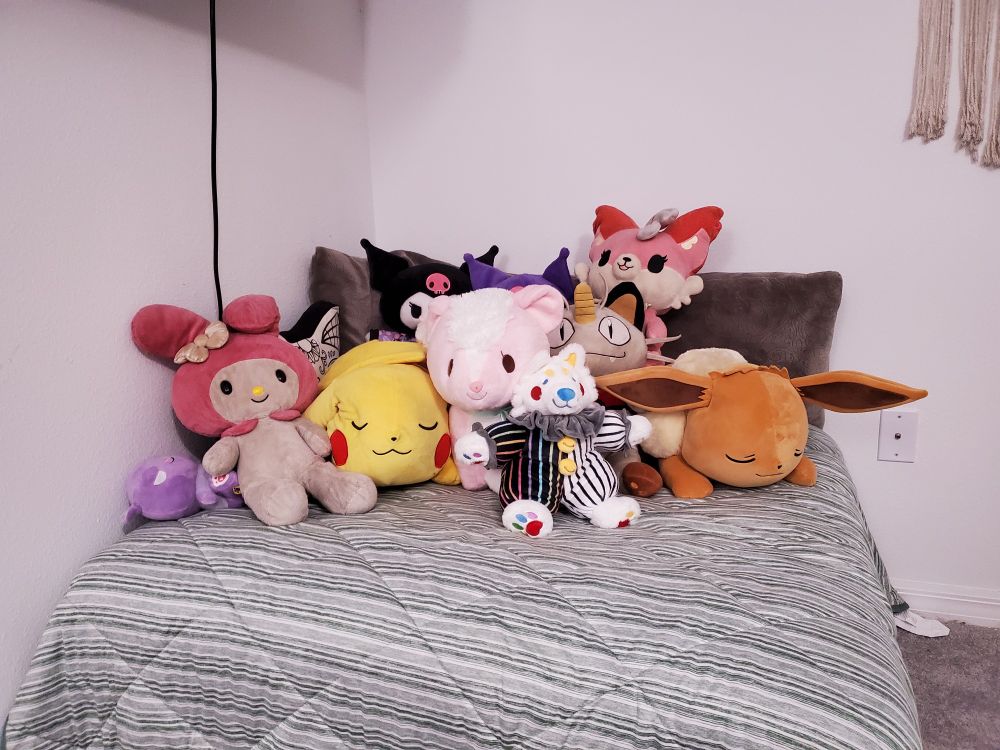 A pile of various stuffed animals on top of a square ottoman nestled into the corner. Sanrio, Pokemon and other colorful plushies all piled up on a striped blanket. 