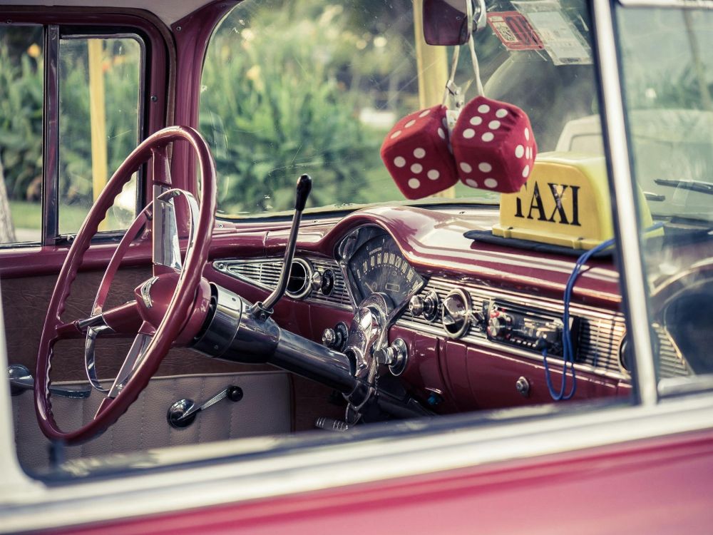 Inside view of the steering column and dash of an old-timey red car. Sign in window says “Taxi” and there are red dice hanging from the rear view mirror.